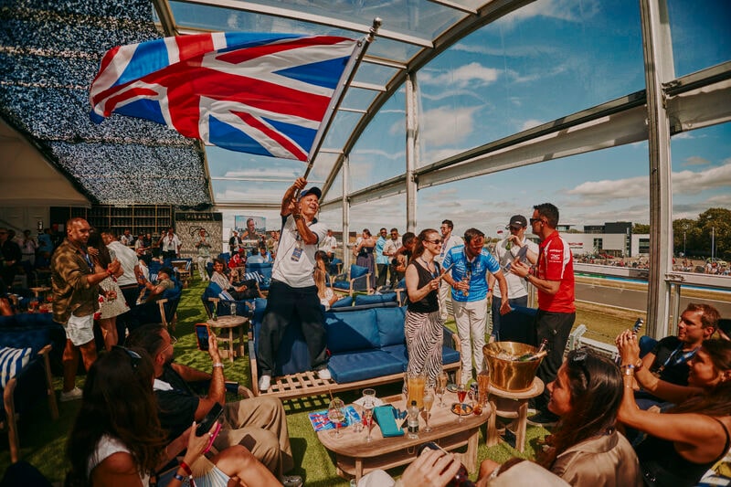 Guests inside the Pira&ntilde;a pavilion at Silverstone, a Union Jack raised over the crowd and the circuit visible through the side of the room