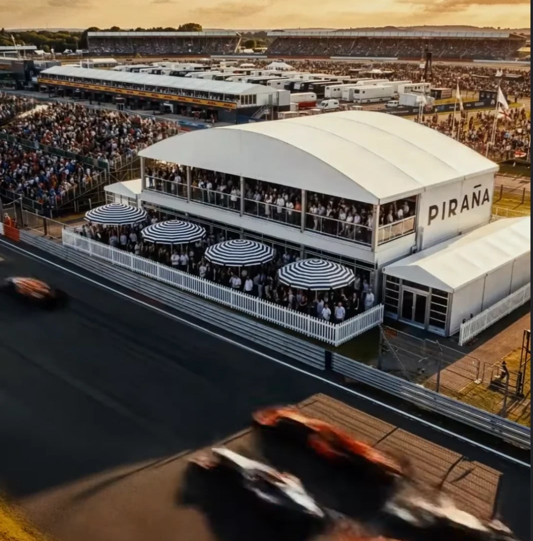 The two-storey Pira&ntilde;a pavilion on the Hangar Straight at Silverstone, terraces full, with Formula 1 cars passing on the track below